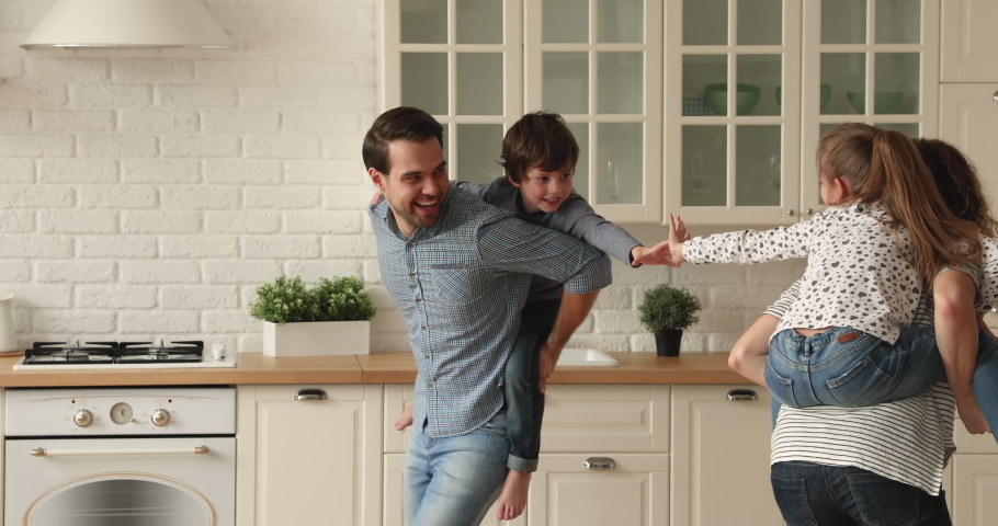 Active young couple piggyback little son and daughter play tag catch me game run at modern kitchen. Parents enjoy funny playtime with cute small kids. Quarantine leisure with children at home concept