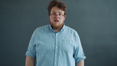 Portrait of guy in glasses sneezing looking at camera standing against gray background alone wearing casual clothing. Disease symptoms and bad health concept.