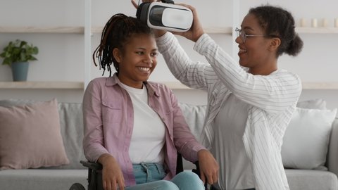 VR Technology For Disabled Person. Happy Handicapped African Teen Girl In Wheelchair Experiencing Augmented Reality, Mother Putting VR Glasses On Daughter's Head At Home. Modern Fun And Entertainment