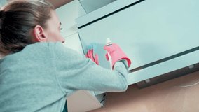 A beautiful young blonde girl washes the kitchen hood with a cleaning agent and a rag. Professional cleaner. Hands in pink rubber gloves.