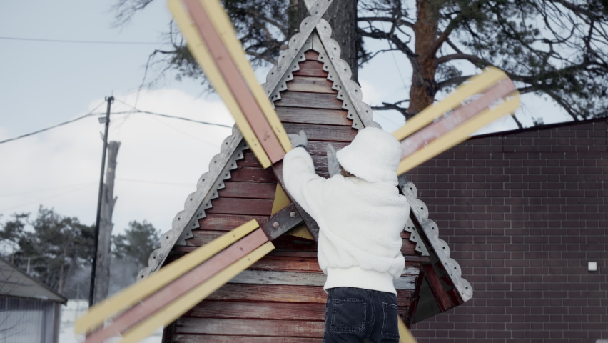 Cheerful teenager girl rotating wooden windmill in winter countryside park. Happy young woman posing on countryside mill on winter walk in village