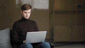 Young focused guy student sitting on couch at home studying remotely with computer during quarantine, e-learning. Caucasian man 25s businessman freelance designer working sitting on sofa using laptop