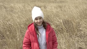 A girl in a red jacket and hat stands against the background of ears of corn and looks into the chamber. The child smiles at the camera while standing in the autumn field.