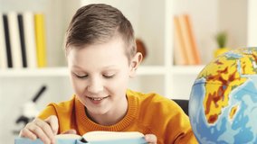 cheerful kid reading book near globe and showing thumb up at home