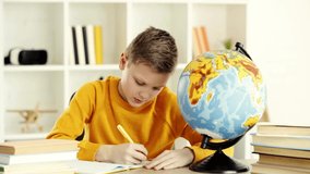 schoolboy writing in notebook and looking at globe while doing homework
