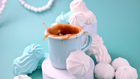 A meringue figurine falls into a cup of black coffee and splashes fly. Espresso with froth and meringues of different colors on a blue background. A creative shot of protein and sugar cookies. 