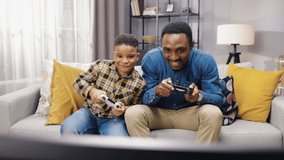 Portrait of African American joyful young male parent having fun with small son sitting on sofa in an apartment and playing video games on a console in good mood. family time, playtime, gamer concept.