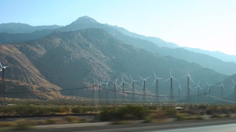 Wind turbine farm in a valley with a mountain range behind on a bright sunny day, next to railway tracks and power lines - California, USA
