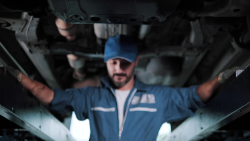 Portrait of a mechanic repairing in uniform standing looking camera at under lifted car of automobile. Repairing car service concept. Royalty-Free Stock Footage #1070395066