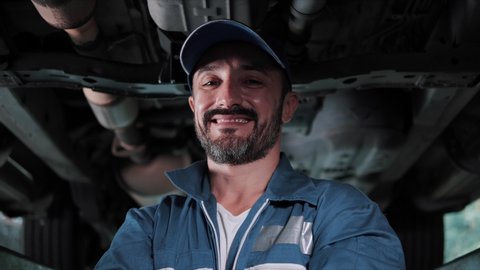Portrait of a mechanic repairing in uniform standing looking camera at under lifted car of automobile. Repairing car service concept.