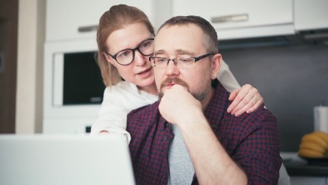 A serious couple husband and wife talking while sitting in the kitchen and looking at a laptop screen. Married spouse checking banking online application on a laptop, receiving bad news notification.