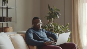 Young Afro-American man in wireless earphones sitting on sofa in living room and speaking via online video call on laptop