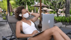 Remote work at the beach during pandemic. Young caucasian woman in protective mask works on a laptop while sunbathing by the pool. 