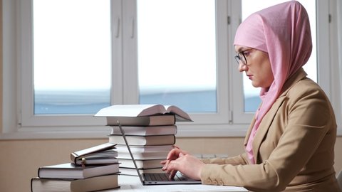 Side view of a young muslim woman in a pink hijab typing on a laptop among the stacks of books.
