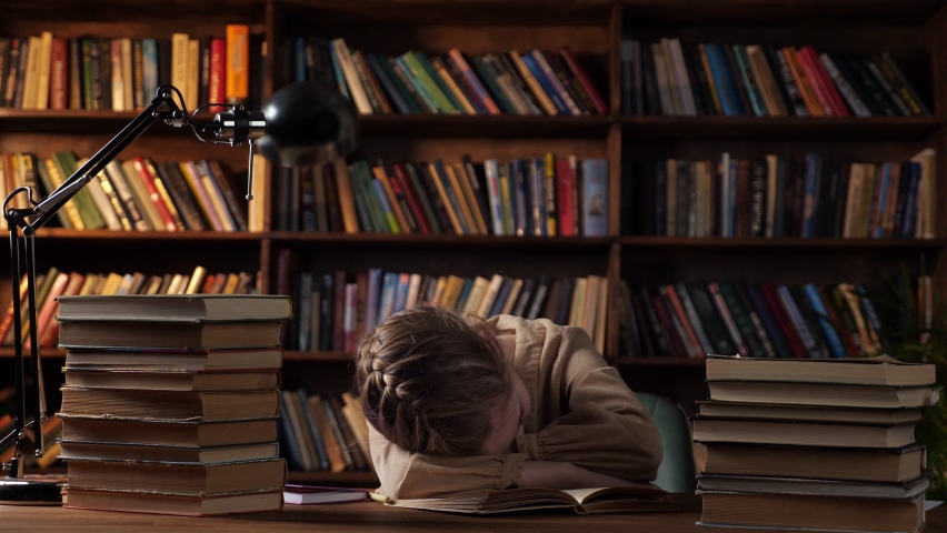 Tired schoolgirl sleeps putting head on copybook among book stacks on wooden table against bookcase under electric light in late evening at home