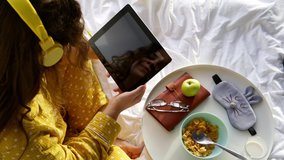 Attractive young woman in pajama uses video chat on tablet near tray with breakfast on comfortable bed in hotel room in lazy morning closeup