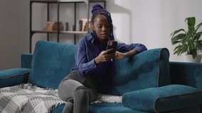 portrait of black teen girl communicating by video chat in smartphone, lady with dreadlocks is sitting on couch in modern apartment