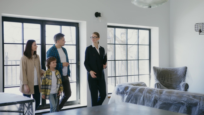 Professional real estate agent welcomes young couple at home showing. Young couple viewing new property and ready to become homeowners. Spacious bright apartment with big windows.4K UHD