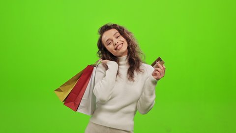 The woman is looking at the camera and smiling. She is holding shopping bags and waving a credit card. She is standing on a green background. Green screen. 4K