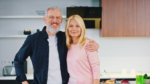 Happy mature couple man and woman stand embracing in the kitchen. Gray bearded husband and blonde wife, stay at home concept