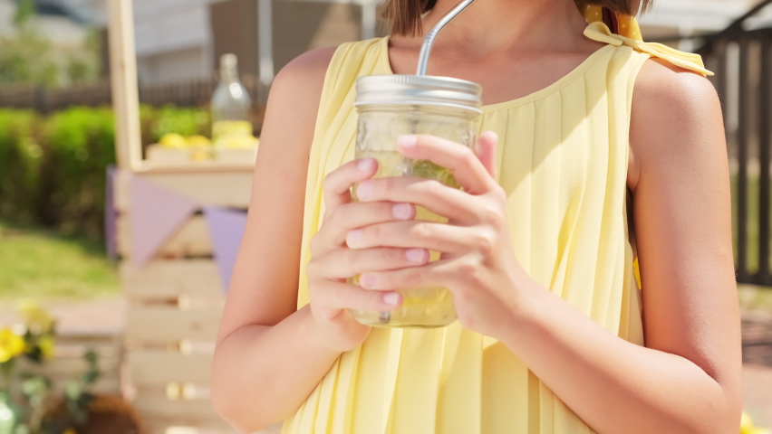 Tilt up closeup of cute pretty 10-year-old girl in cool summer hat drinking freshly made lemonade and smiling to camera standing outdoors on summer day