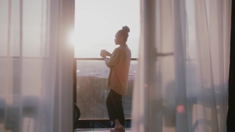 Shot of young african american woman stand on balcony looking out over city drinking a cup of tea or coffee. Relaxation,travel, interior, apartment. Slow motion