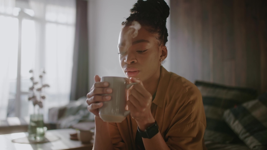 Young beautiful african woman drink cup of coffee or tea sitting at home. Thinking, holding, inspiration, dreaming, cute, positive, relaxation. Close up. Slow motion Royalty-Free Stock Footage #1070421019