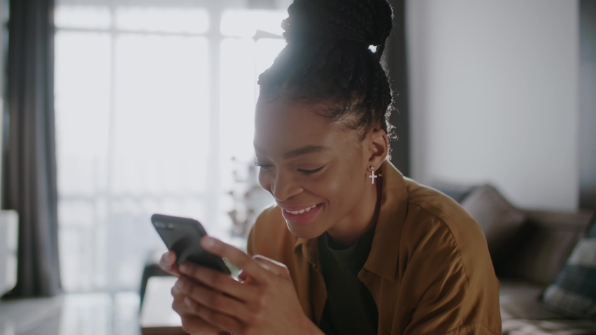 Smiling afro american woman hold use smart phone sitting at home at sunlight. Watching social media. Portrait, entertainment, smartphone. Slow motion Royalty-Free Stock Footage #1070421052