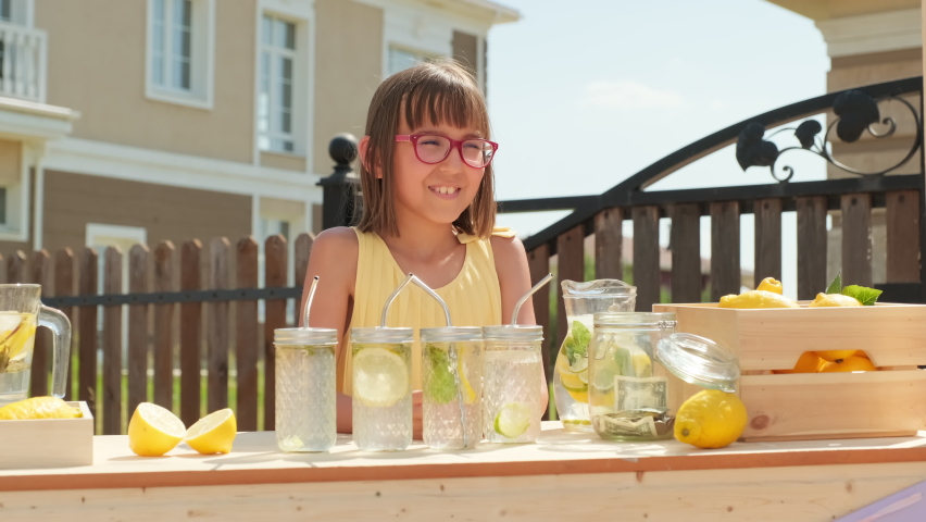 Waist-up shot of cheerful 10-year-old girl selling fresh homemade lemonade to woman standing outdoors at diy stand on hot summer day