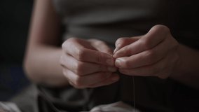 4K Close-up view of female hands inserting a thread into a needle hole for sewing. Young woman in green dress doing needlework. Hand made concept. Daylight.