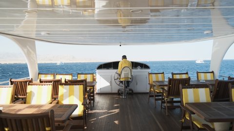 Captain of cruise yacht controls boat, side view. Senior man in yellow uniform turns steering wheel of ship in sunny windy day. White yacht sailing on blue sea. Empty chairs for tourists on deck