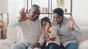 Afro american happy family sitting at home sofa waving hello looking at camera, little girl daughter black child showing cool gesture two fingers, ethnic african parents with baby kid greeting indoor