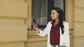 Cheerful female manager having phone conversation on urban street. Smiling mixed race woman speaking on video call outside. Confident businesswoman walking with smartphone outdoors.