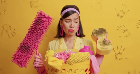 Puzzled frustrated housewife looks at dirty mop and sponge busy doing house chores doesnt like dirt stands near basket of laundry isolated over yellow background. Domestic responsibilities concept