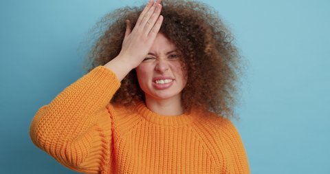 Dissatisfied curly haired woman keeps hand on forehead regrets doing something wrong shakes head looks confused dressed in casual orange sweater isolated over blue background. Negative emotions