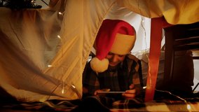 Little boy in Santa hat in DIY kid's tent laughing celebrating Christmas using smartphone, having joyful online conversation. Christmas fun at home.