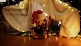 Happy little boy in Santa hat lying in DIY child tent celebrating Christmas using smartphone, having video call. Christmas night, home entertainment, long distance communication concept.