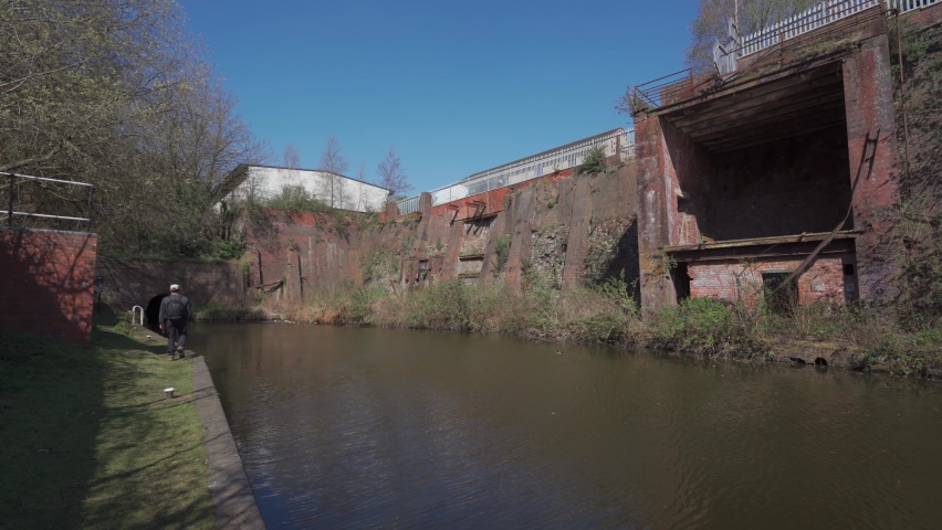 A man walking along a canal past old derelict industrial brickwork remains