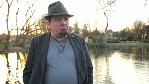 [4] dramatic portrait of goatee man wearing hat in angry standing in front of pond in sunset