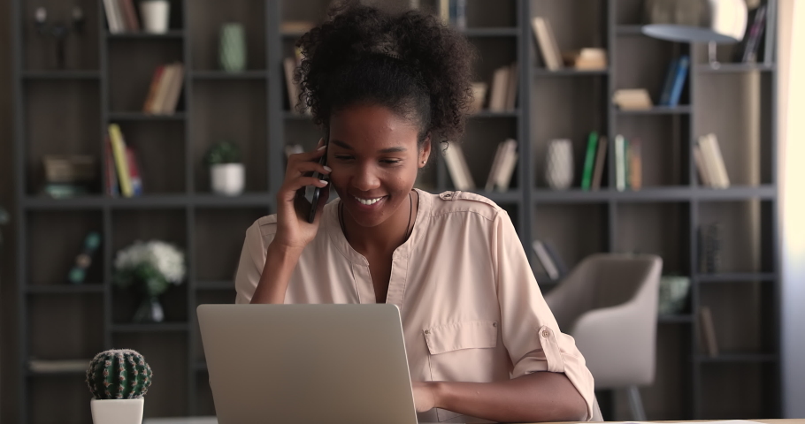Focused millennial african american business lady working on computer, giving consultation in cellphone conversation to client or discussing project issues with colleagues distantly, multitask concept