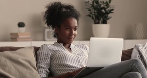Relaxed millennial smiling african mixed race woman looking at laptop screen, watching funny movie film or pleasant video photo content in social network, shopping in internet store or web surfing.