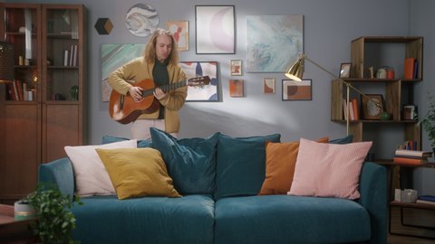 Happy man playing acoustic guitar falling and lying on sofa, singing song. Creative hobby. Rock crazy musician enjoying his music instrument, having fun at home. 
