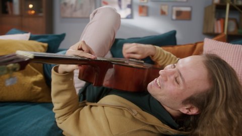 Happy man playing acoustic guitar lying on sofa, singing song. Creative hobby. Rock crazy musician enjoying his music instrument, having fun at home. 