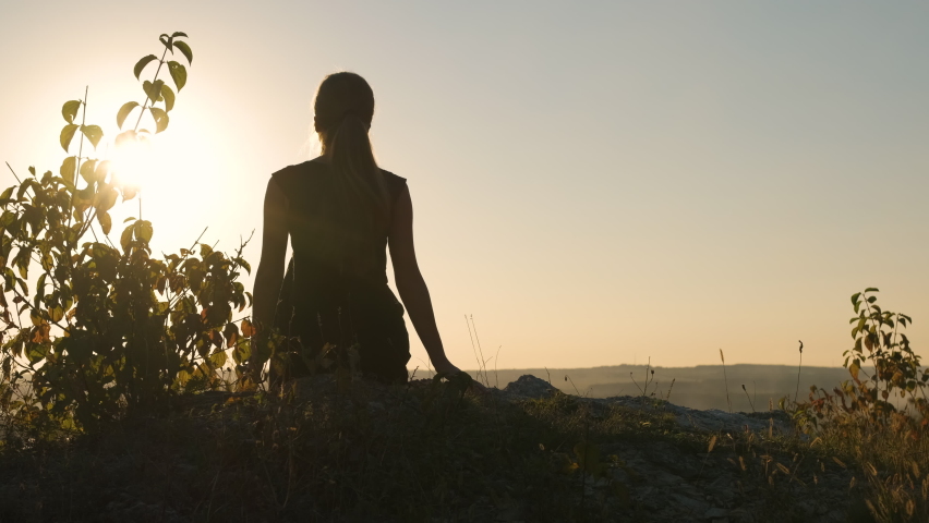 Young elegant woman in black short dress and white sneaker shoes sitting on a rock relaxing outdoors at summer evening. Fashionable lady enjoying warm sunset in nature.