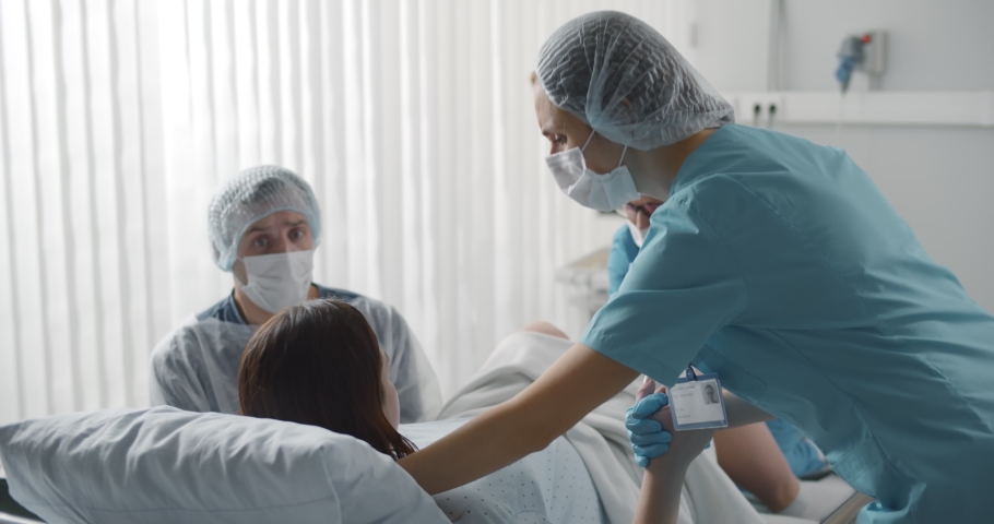Woman giving birth with husband holds her hand in support and obstetricians assisting. Back view of medical staff in protective uniform helping pregnant woman in labor Royalty-Free Stock Footage #1070475619