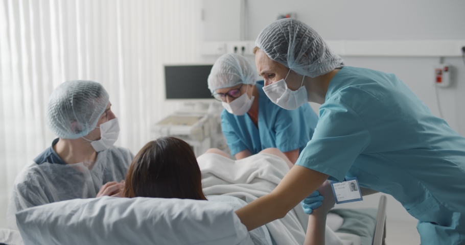 Woman giving birth with husband holds her hand in support and obstetricians assisting. Back view of medical staff in protective uniform helping pregnant woman in labor | Shutterstock HD Video #1070475619