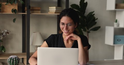 Smiling young beautiful businesswoman in eyewear enjoying working on computer, communicating distantly with partners or client, using laptop software applications sitting at desk at home or office.