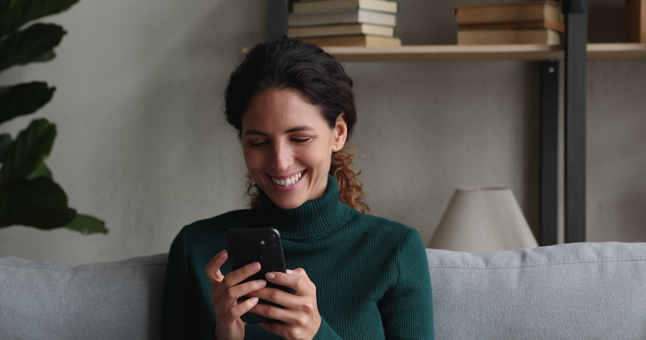 Happy millennial beautiful woman looking at mobile phone screen watching funny photo or video, enjoying pleasant distant communication in social network, web surfing or reading good news at home. Royalty-Free Stock Footage #1070487955