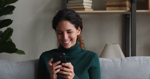 Happy millennial beautiful woman looking at mobile phone screen watching funny photo or video, enjoying pleasant distant communication in social network, web surfing or reading good news at home.