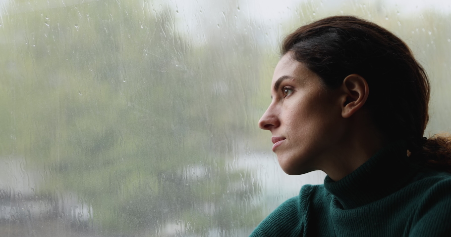 Close up head shot melancholic lost in thoughts young woman sitting near window, looking outside contemplating rainy gloomy weather, recollecting memories, dreaming or visualizing future alone indoors | Shutterstock HD Video #1070487970
